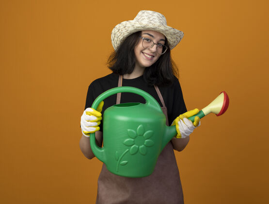姿势微笑着的年轻黑发女园丁戴着眼镜 穿着制服 戴着园艺帽和手套 拿着隔离在橙色墙上的浇水罐花园眼镜手套