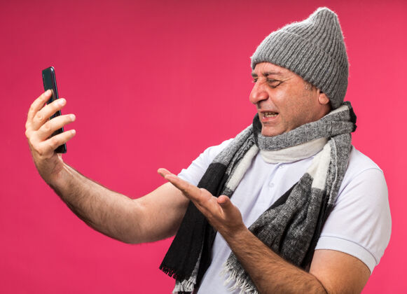 男人恼怒的成年白种人 脖子上围着围巾 戴着冬天的帽子 拿着手机 指着粉色墙壁上的电话 还有复印空间围巾孤立戴着