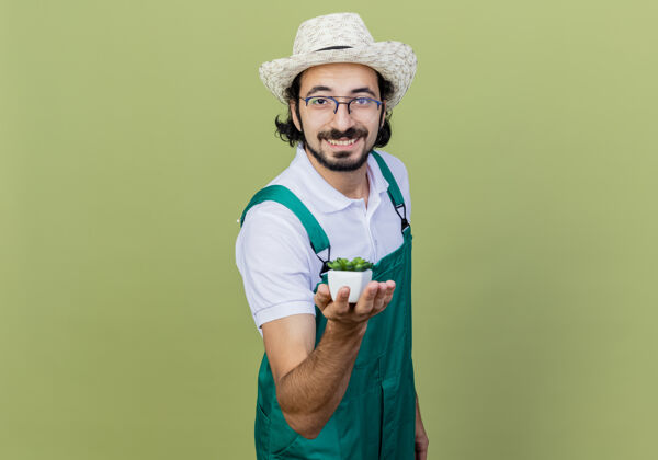 感觉年轻的留着胡须的园丁 穿着连身衣 戴着帽子 展示着盆栽植物 站在浅绿色的墙上 面带微笑地看着前方衣服快乐脸