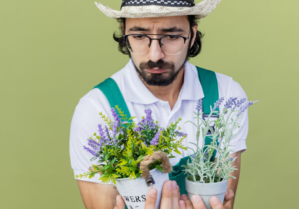 人年轻的留着胡子的园丁 穿着连体衣 戴着帽子 手里拿着盆栽植物 站在浅绿色的墙上 带着悲伤的表情看着它们连身衣人胡子