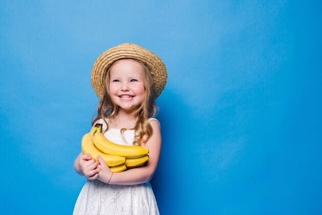 漂亮快乐的小女孩和黄色的香蕉隔离在蓝色的墙上白裙子女孩香蕉