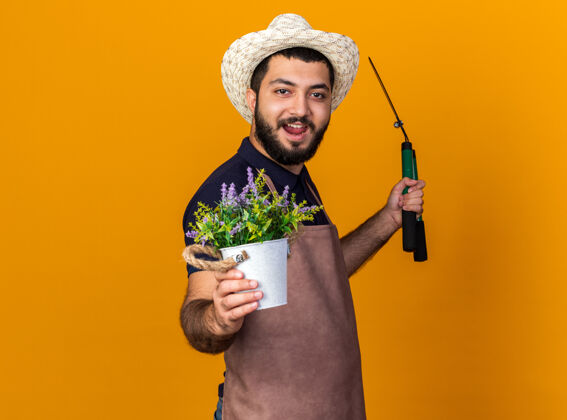 侧快乐的年轻白人男性园丁戴着园艺帽站在一旁 手持园艺剪刀和花盆 隔离在橙色的墙上 留有复制空间花园白种人立场
