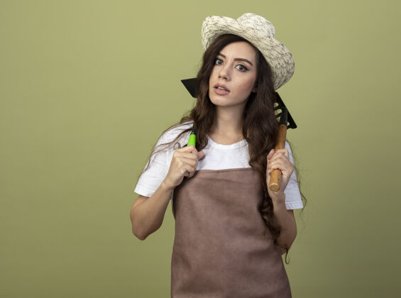 肩穿着制服 戴着园艺帽的焦虑的年轻女园丁把耙子和锄头耙子放在隔离在橄榄绿墙上的肩膀上制服园艺耙子