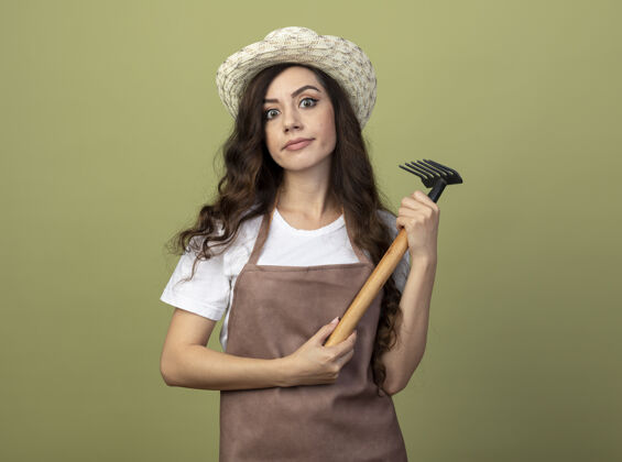 帽子年轻的女园丁穿着制服 戴着园艺帽 拿着耙子 望着橄榄绿墙上孤立的前方脸印象耙子