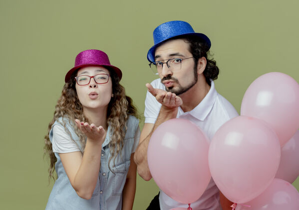 亲吻高兴的年轻夫妇戴着粉红和蓝色的帽子站在气球后面 并显示亲吻的手势气球表演粉色