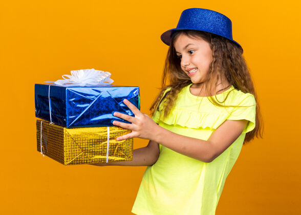 帽子兴奋的白人小女孩拿着蓝色派对帽 看着橙色墙上的礼品盒 还有复印空间小兴奋抱着