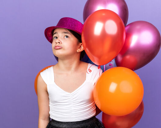 帽子戴着紫色派对帽 手持氦气球 孤零零地仰望着紫色墙壁 留着复制空间的悲伤白人女孩气球年轻孤立