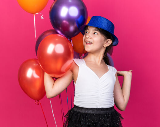 站快乐的年轻白人女孩 戴着蓝色派对帽 站在氦气球前 看着粉色墙壁上的孤立复制空间帽子女孩氦