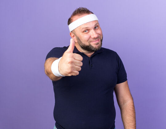 拇指高兴的成年斯拉夫运动男子戴着头带和腕带竖起大拇指孤立的紫色墙壁与复制空间成人拜托孤立型