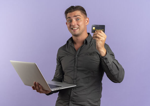 笔记本电脑年轻惊讶的金发帅哥拿着笔记本电脑和信用卡背景紫罗兰持有