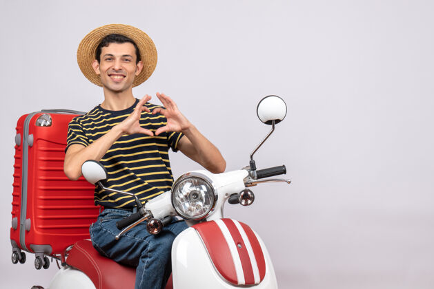 微笑骑在轻便摩托车上戴草帽的年轻人的正视图 正在做心脏标志摩托车成人输送机