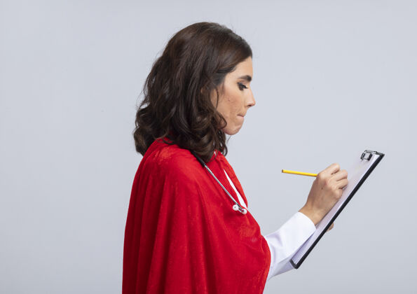 脸自信的女超人穿着医生制服 戴着红色斗篷和听诊器 站在一边 用铅笔在剪贴板上写字 隔离在白墙上听诊器表情医生