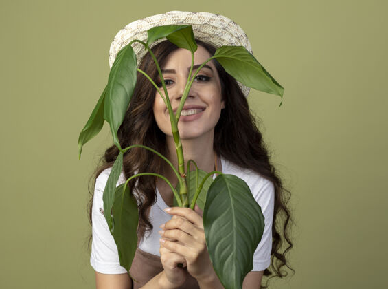 制服身着制服 戴着园艺帽 面带微笑的年轻女园丁将植物孤立在橄榄绿的墙上女人穿着前面