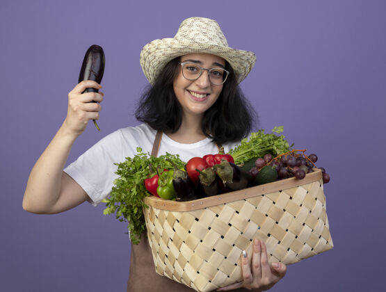 人年轻的深色皮肤的女园丁微笑着 戴着眼镜 穿着制服 戴着园艺帽 把菜篮子和茄子隔离在紫色的墙上脸市民女性