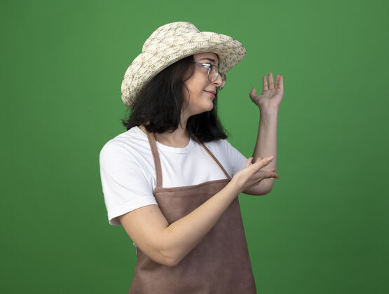 眼镜高兴的是年轻的黑发女园丁戴着眼镜和制服 戴着园艺帽 双手孤立在绿色的墙上手市民背部