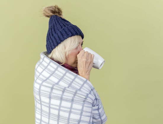 人自信的年轻金发病女人戴着冬天的帽子 围巾裹着格子呢 站在一旁喝着橄榄绿墙上孤立的杯子表情围巾斯拉夫