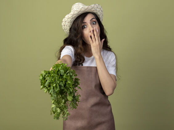 人身着制服 头戴园艺帽的年轻女园丁惊讶地把手放在嘴上 把香菜隔离在橄榄绿的墙上女人人脸