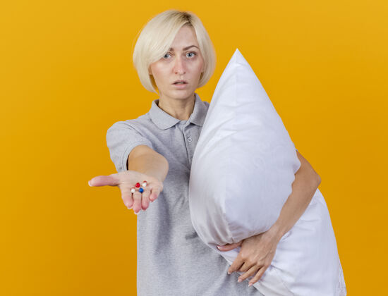 金发自信的年轻金发病女人抱着枕头 手里拿着隔离在橙色墙上的药片表情自信枕头