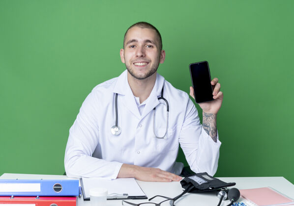 医疗面带微笑的年轻男医生穿着医用长袍和听诊器坐在办公桌旁 手里拿着工作工具 手放在桌子上 隔离在绿色的墙上工具穿着表演