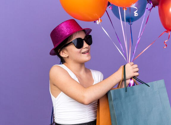 年轻戴着太阳眼镜 戴着紫色派对帽 手里拿着购物袋 面带微笑的白种人小女孩 看着隔离在紫色墙上 留有复制空间的氦气球氦女孩包