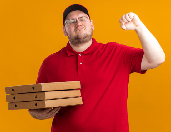 披萨自信超重的年轻送货员戴着眼镜 手里拿着披萨盒 手举拳头 站在橙色的墙上 留着复印空间拳头送货员孤立