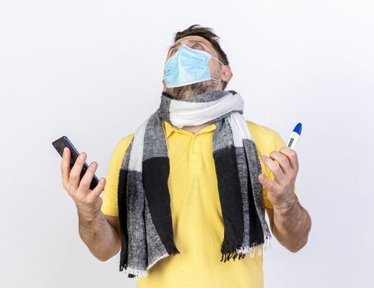 惊讶一个戴着医用面罩和围巾的年轻金发病患把电话和温度计隔离在白墙上围巾斯拉夫面具