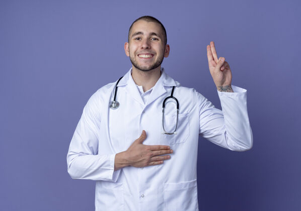 人面带微笑的年轻男医生穿着医用长袍和听诊器做承诺手势隔离在紫色的墙上男性承诺姿势
