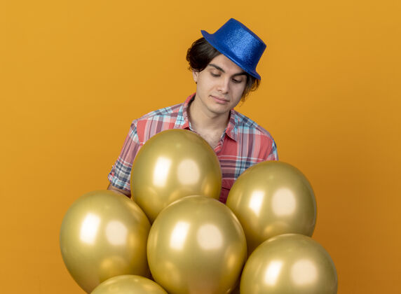 站戴着蓝色派对帽的帅哥高兴地看着站在橙色墙上的氦气球人气球请