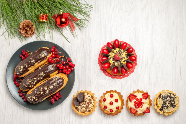 圣诞节俯视图：灰色平板上的巧克力和醋栗 底部的浆果蛋糕馅饼 白色木质地面上的松树叶子和圣诞玩具 还有复制空间蛋糕花馅饼