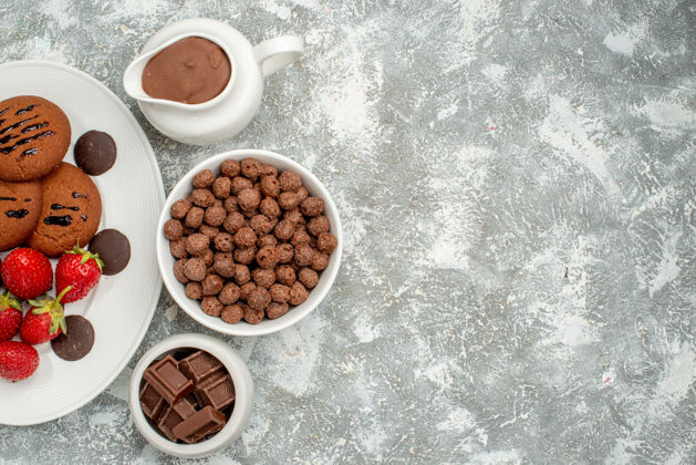 碗上半部分是白色椭圆形盘子上的巧克力饼干草莓和圆形巧克力 灰白色地面上的碗里有巧克力麦片和可可杯子咖啡磨碎的