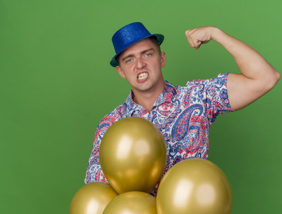 年轻愤怒的年轻人戴着蓝色的帽子站在气球后面 表现出强烈的姿态孤立在绿色帽子气球绿色