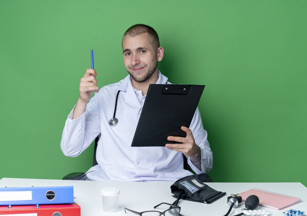 穿着年轻的男医生穿着医用长袍 手持听诊器 坐在办公桌旁 手里拿着写字板和笔 隔离在绿色的墙上人感觉衣服