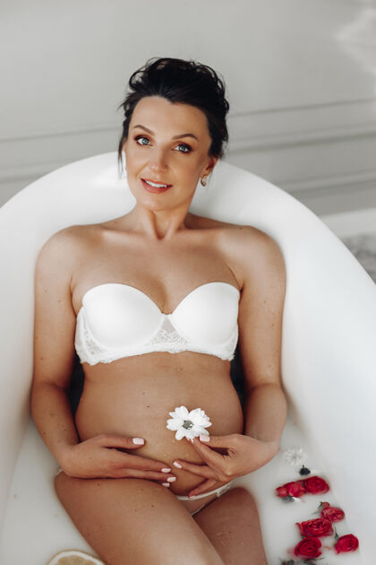 期望有吸引力的怀孕成人的肖像 深色头发 穿着白色胸罩 躺在浴缸里 肚脐上开着白色的花 水里放着红玫瑰孕妇微笑着休闲女性女人