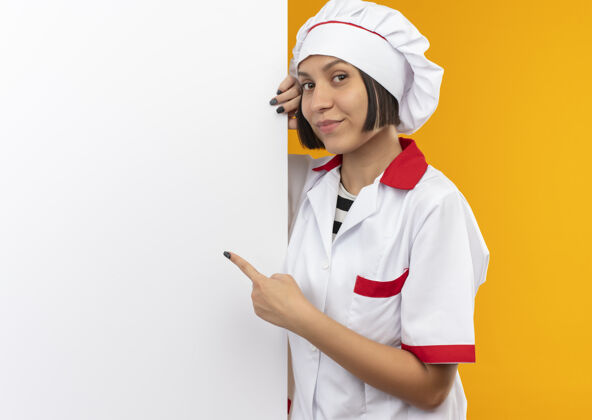 表情身着厨师制服的年轻女厨师高兴地站在身后 指着隔离在墙上的白色墙壁请脸人