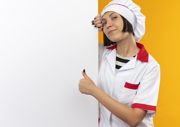 请身着厨师制服的年轻女厨师站在白色的墙后 孤立地在墙上竖起大拇指衣服拇指白