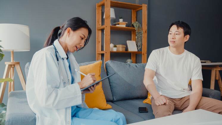 听诊器年轻的亚洲女专业医师医生用数字平板电脑和快乐的男病人分享健康测试的好消息坐在家里的沙发问诊所客厅