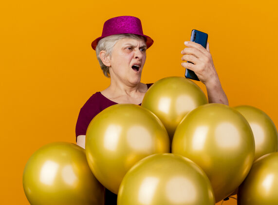 帽子恼怒的老妇人戴着聚会帽站在那里 手里拿着氦气球 看着隔离在橙色墙上的电话人氦老人