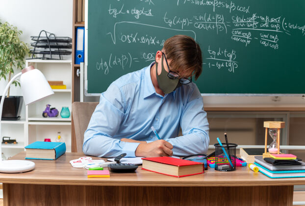 眼镜年轻的男老师戴着眼镜和面罩备课看起来很自信坐在课桌旁 教室里的黑板前放着书和笔记保护老师准备
