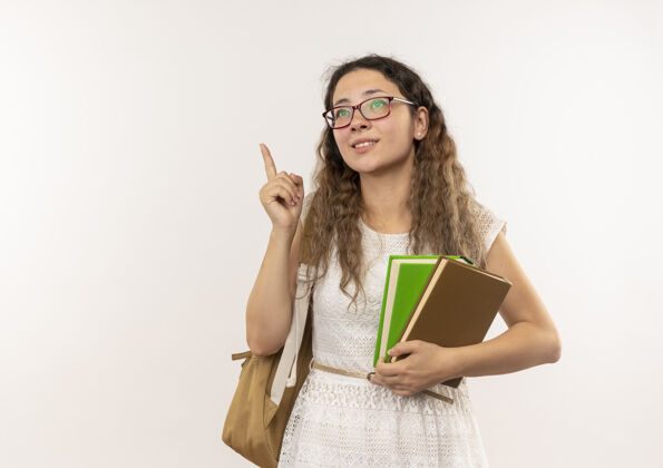 漂亮高兴的年轻漂亮的女学生戴着眼镜 背着书包 抬头抬起手指 孤立地站在白色的墙上手指衣服抱