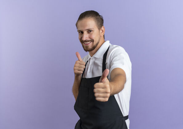 衣服微笑着的年轻英俊的理发师穿着制服 在紫色的墙上孤立的前面竖起大拇指姿势人表演