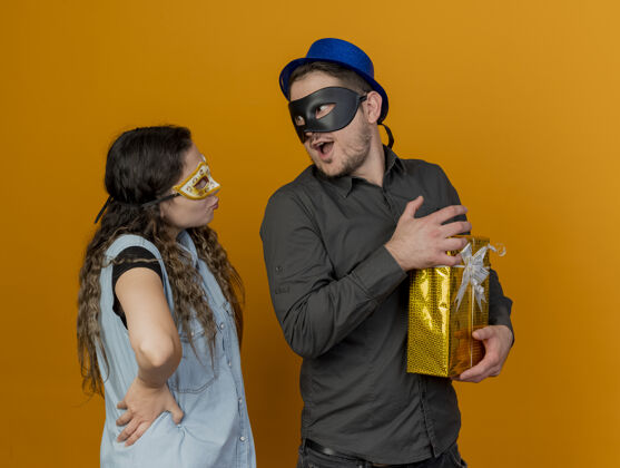 派对年轻的派对情侣戴着化装眼罩看着对方手里拿着礼物的家伙孤立在橙色上面具礼物举行