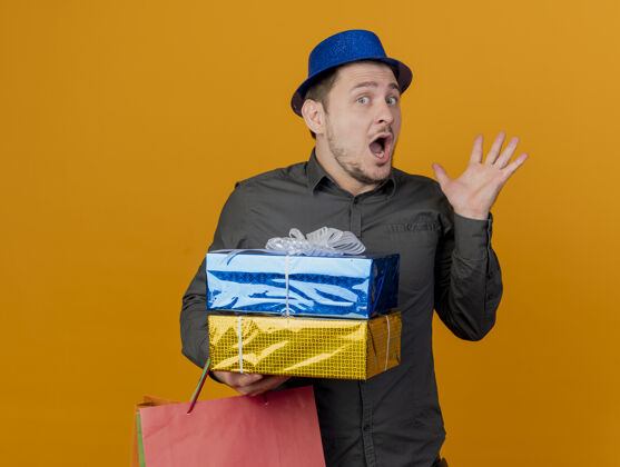 包害怕的年轻人戴着蓝色的帽子拿着礼品盒和包展开手孤立在橙色小伙子橙色盒子