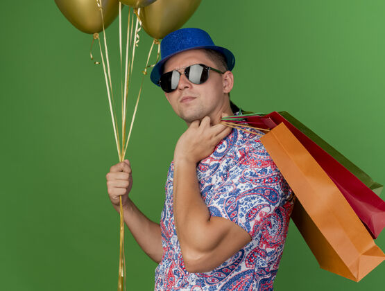 年轻人自信的年轻人戴着蓝色的帽子和眼镜拿着气球 把礼品袋放在肩膀上的绿色隔离礼物小伙子包