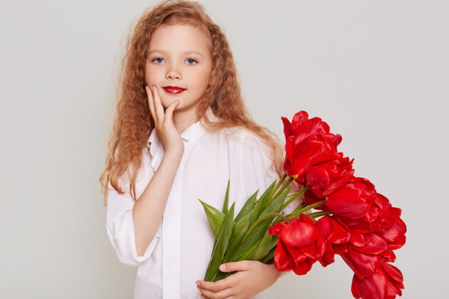 乐趣可爱的小女孩穿着白色衣服拿着一大束红色郁金香作为礼物 看着面前风趣自信的表情笑脸女学生孩子