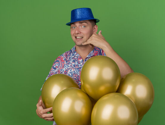 年轻人微笑着的年轻人戴着蓝色帽子站在气球后面 在绿色的舞台上展示着电话的手势电话手势站着