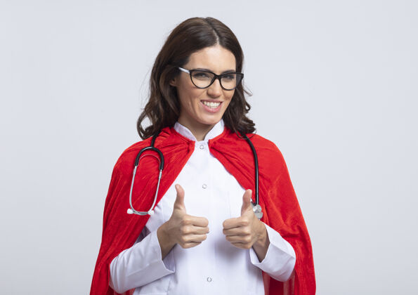 听诊器微笑的女超人穿着医生制服 戴着红色斗篷 听诊器戴着光学眼镜 两只手竖起大拇指孤立在白色的墙上手女孩人