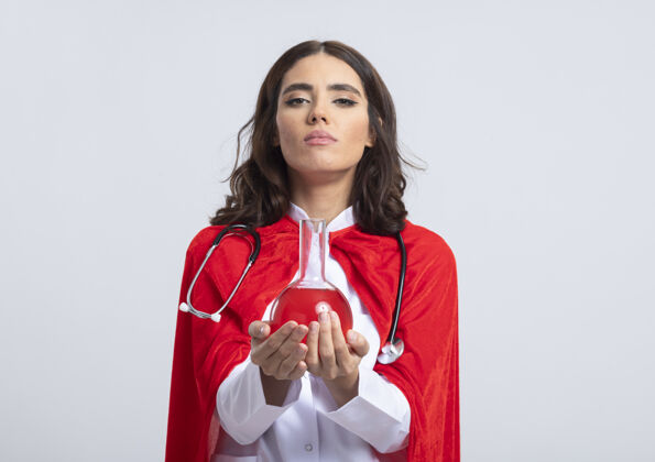 玻璃自信的女超人穿着医生制服 戴着红色斗篷和听诊器 拿着隔离在白墙上的玻璃瓶中的红色化学液体信心人斗篷