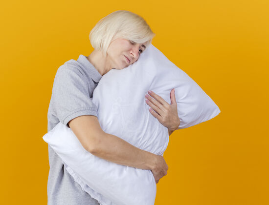 斯拉夫人悲伤的年轻金发病女人抱着枕头看着橙色墙上孤立的一面姿势公民疾病