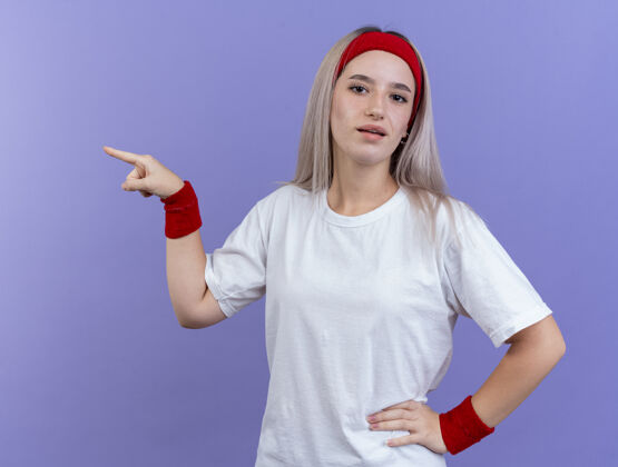 女孩自信的年轻运动女性戴着背带 戴着头带和腕带 指着隔离在紫色墙上的一侧女人年轻人
