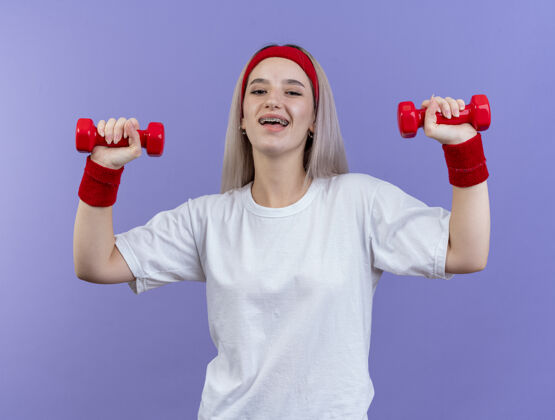 手快乐的年轻运动女性戴着背带 戴着头带和腕带 双手举着哑铃站在紫色的墙上举起腕带快乐